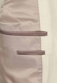  Lichtbeige stof met een gladde textuur, voorzien van twee verticale, bruine ritszakken die aan de zijkant zijn genaaid. Subtiele stikdetails zijn zichtbaar.