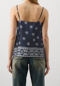 Mörkblå ärmlös topp med vita blommönster och geometriska mönster, med justerbara band och strukturerad nederkant. Matchas med ljusa denimjeans.