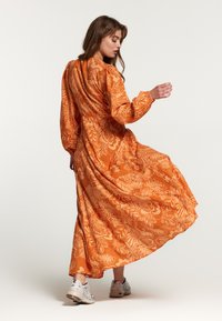 Lang oranje jurk met bloempatroon, lange mouwen, verzamelde rug en vloeiende silhouet. Gecombineerd met witte sneakers.