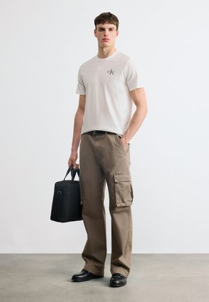 Νεαρός άνδρας στέκεται φορώντας λευκό t-shirt Calvin Klein, καφέ παντελόνι cargo, μαύρα παπούτσια, κρατώντας μια μαύρη τσάντα γραφείου με το ένα χέρι.