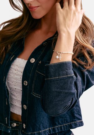 Frau trägt eine dunkle Jeansjacke und ein weißes Spitzenoberteil mit einem Perlenarmband, das ein Kreuz- und Perlenanhänger an ihrem Handgelenk hat.