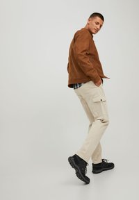 Giacca in suede marrone, pantaloni cargo beige con tasca laterale e scarpe sportive nere. Tessuti a contatto stretto e materiali testurizzati con elementi di design pratici.