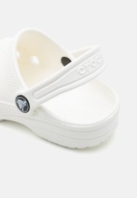 Beli Crocs sandali z teksturirano površino, nastavljivo zanko in krožno detajlno logotip. Lahka struktura in odprt dizajn za prste.
