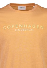 Camiseta de manga corta, de corte relajado, en naranja claro con un degradado. Presenta un texto gráfico en blanco y amarillo que dice "COPENHAGUE" y "LINDBERGH."