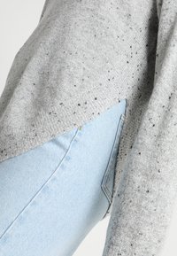 Grau gesprenkelter Strickpullover mit lockerer Passform, kombiniert mit hellblauen Denim-Jeans mit einer seitlichen Tasche und einem leichten Verblassen.