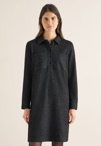 Donna con lunghi capelli castani che indossa un vestito nero a maniche lunghe con bottoni e motivo a spina di pesce, su sfondo beige.