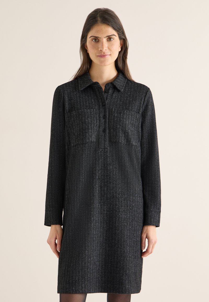 Donna con lunghi capelli castani che indossa un vestito nero a maniche lunghe con bottoni e motivo a spina di pesce, su sfondo beige.