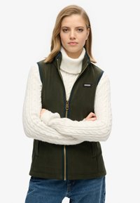 Ciemnozielona kamizelka z polarowego materiału z zamkiem, granatowymi wykończeniami i bocznymi kieszeniami, noszona na białym swetrze z tkaniny w warkocze z golfem.