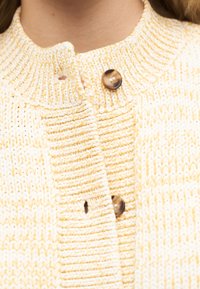 Κοντινό πλάνο σε πλεκτό πουλόβερ σε κρέμ και κίτρινο χρώμα με δύο καφέ κουμπιά κλεισμένα κοντά στον γιακά.