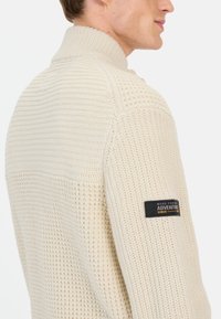 Strikket sweater i creme med høj krave, prydet med teksturerede mønstre og et sort patchmærke på den venstre ærme.
