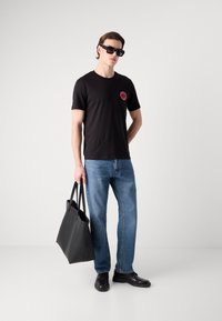 Black t-shirt with a red circular logo, blue jeans, and black leather shoes. Holding a large black leather tote bag. Sunglasses worn.