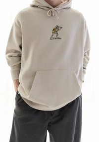 Beżowa bluza z kapturem, z przednią kieszenią, sznurkiem do regulacji kaptura oraz haftowanym obrazkiem postaci trzymającej tarczę i tekstem poniżej.