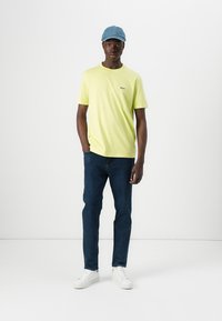 Lys gul kortermet t-skjorte med svart brystlogo, kombinert med mørkeblå jeans og hvite sneakers. Tilbehør med en lys blå caps.