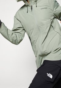 Giacca impermeabile verde chiaro con zip frontale, cappuccio regolabile e dettagli neri. Abbinata a pantaloni neri con tasca brandizzata.