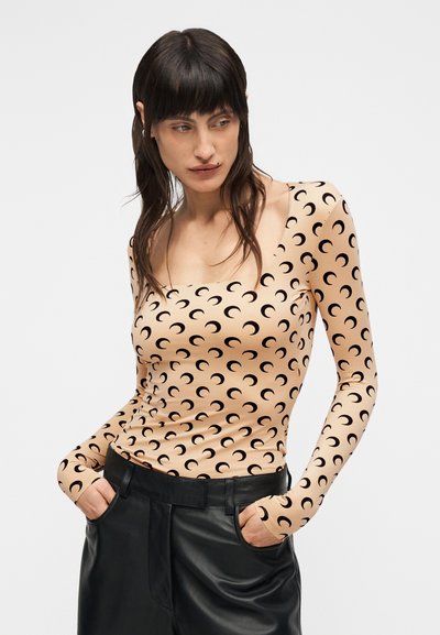 Μπλούζα με μακριά μανίκια σε ανοιχτό μπεζ ύφασμα με σχέδιο μαύρου ημισελήνου. Διαθέτει τετράγωνη λαιμόκοψη και εφαρμοστή σχεδίαση, συνδυασμένη με μαύρο παντελόνι.