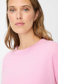 Maglione rosa chiaro con collo rotondo, trama morbida e design minimalista; indossato da una persona con capelli lunghi e ondulati e trucco naturale.