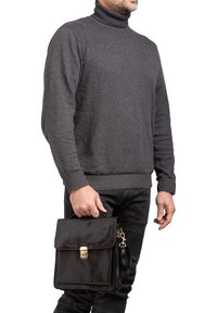 Borsa a tracolla nera in pelle con patta, chiusura dorata e dettagli con zip. Il modello indossa un dolcevita grigio e jeans neri.