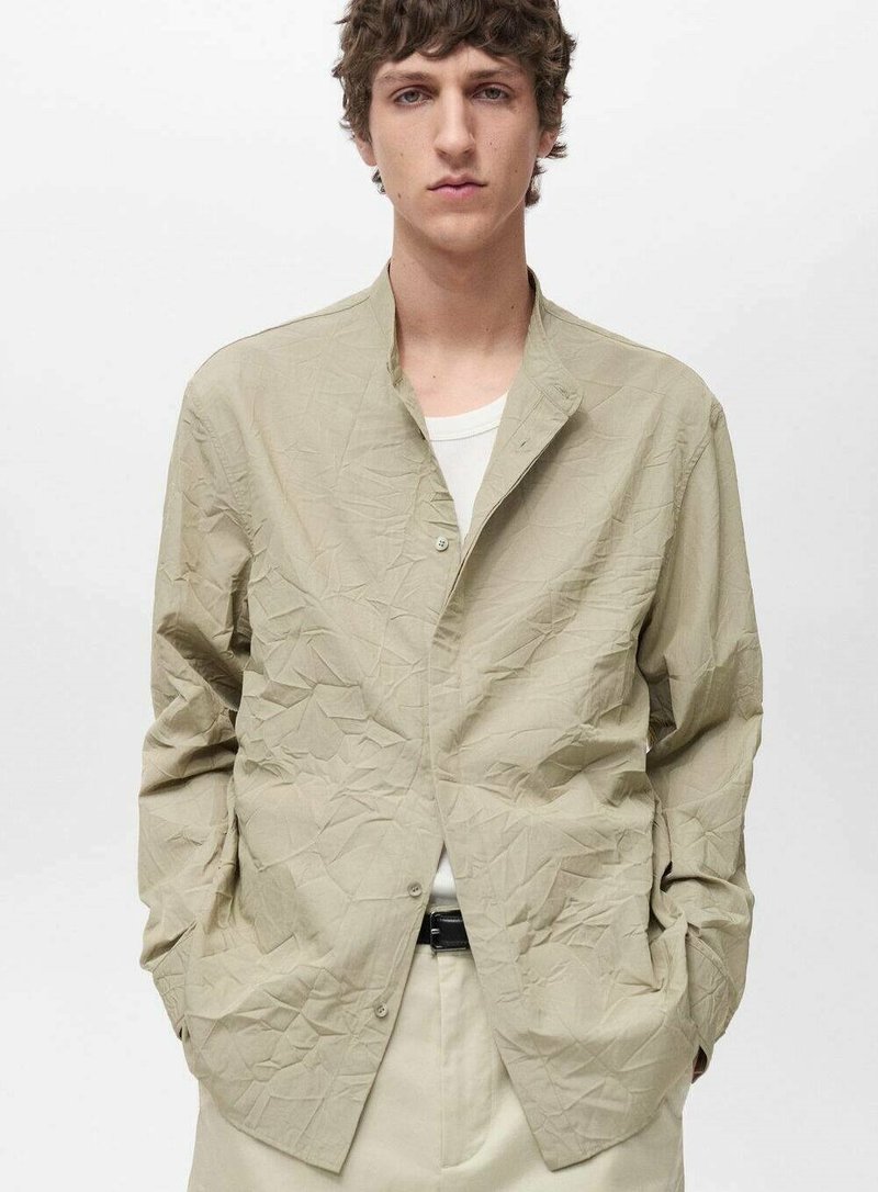 Camicia di stoffa beige, stropicciata, con colletto rilassato e chiusura a bottoni. Maniche lunghe, vestibilità ampia, abbinata a una maglietta bianca sotto.