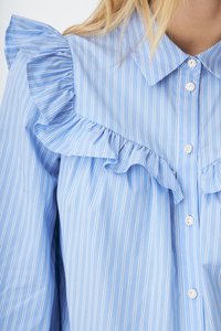 Kvinne iført en lyseblå skjorte med vertikale striper, med rysjedetaljer på skuldrene og brystet, og dekorative perleknapper.