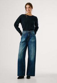 Una mujer joven está de pie con las manos en los bolsillos, vistiendo un suéter de punto negro, jeans de tiro alto de mezclilla azul desgastada y zapatos de punta negros sobre un fondo liso.