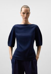Marinblus med bred båtutskärning och korta, puffiga ärmar. Tillverkad av ett smidigt tyg, har en figursydd silhuett och en subtil glans.