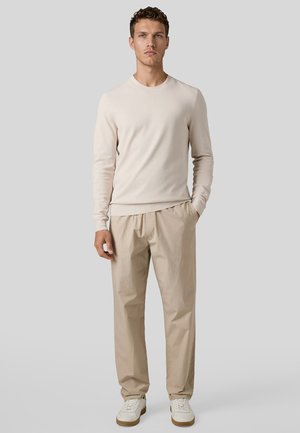 Ung mand stående iført en beige langærmet skjorte, lysebrune bukser og hvide sneakers mod en ensartet lysegrå baggrund.
