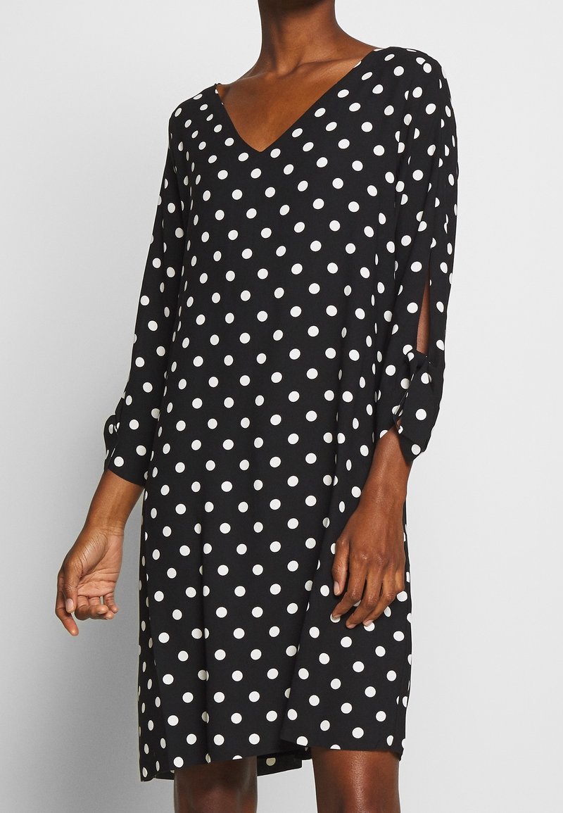 Musta polkapilkkumekko, jossa valkoiset pilkut, V-kaula-aukko, pitkät hihat halkioilla ja rento siluetti. Pehmeä kangas, jossa sileä pinta.