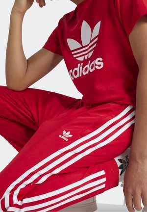 Person trägt roten Adidas-Trainingsanzug mit weißen drei Streifen, Adidas-Logo auf Shirt und Hose, sitzt mit der Hand auf dem Knie ruhend.
