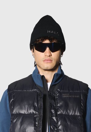 Zwarte gebreide muts, reflecterende zonnebril en een donker puffer vest met blauwe fleece accenten en logo details op de borst.