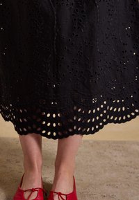 Lower legs wearing red shoes under a black skirt with embroidered floral patterns and scalloped hem with cut-out details.