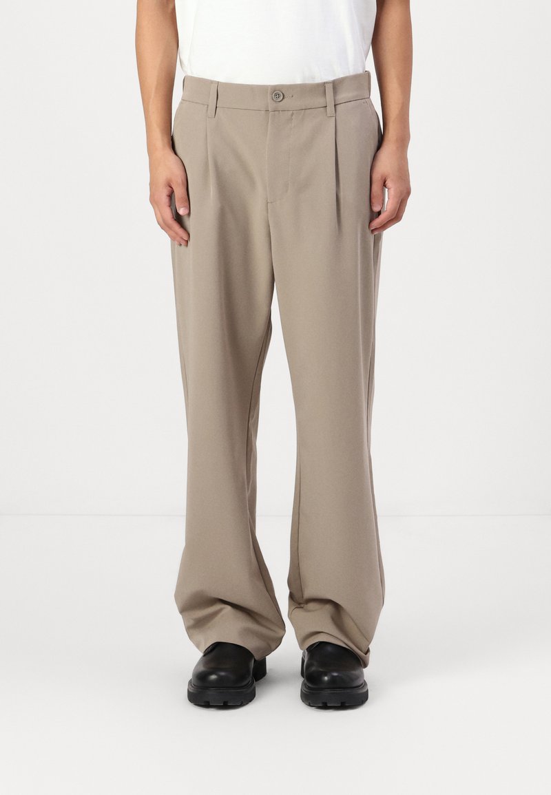 Beige wide-leg trousers in a smooth fabric with front pleats, a single button closure, and belt loops, paired with black sturdy footwear.