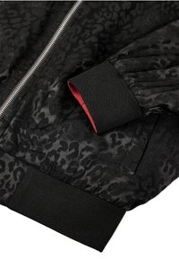 Czarna kurtka typu bomber z teksturowanym wzorem w cętki, srebrnym zamkiem błyskawicznym, ściągaczami z czerwonymi akcentami i kieszeniami po bokach.