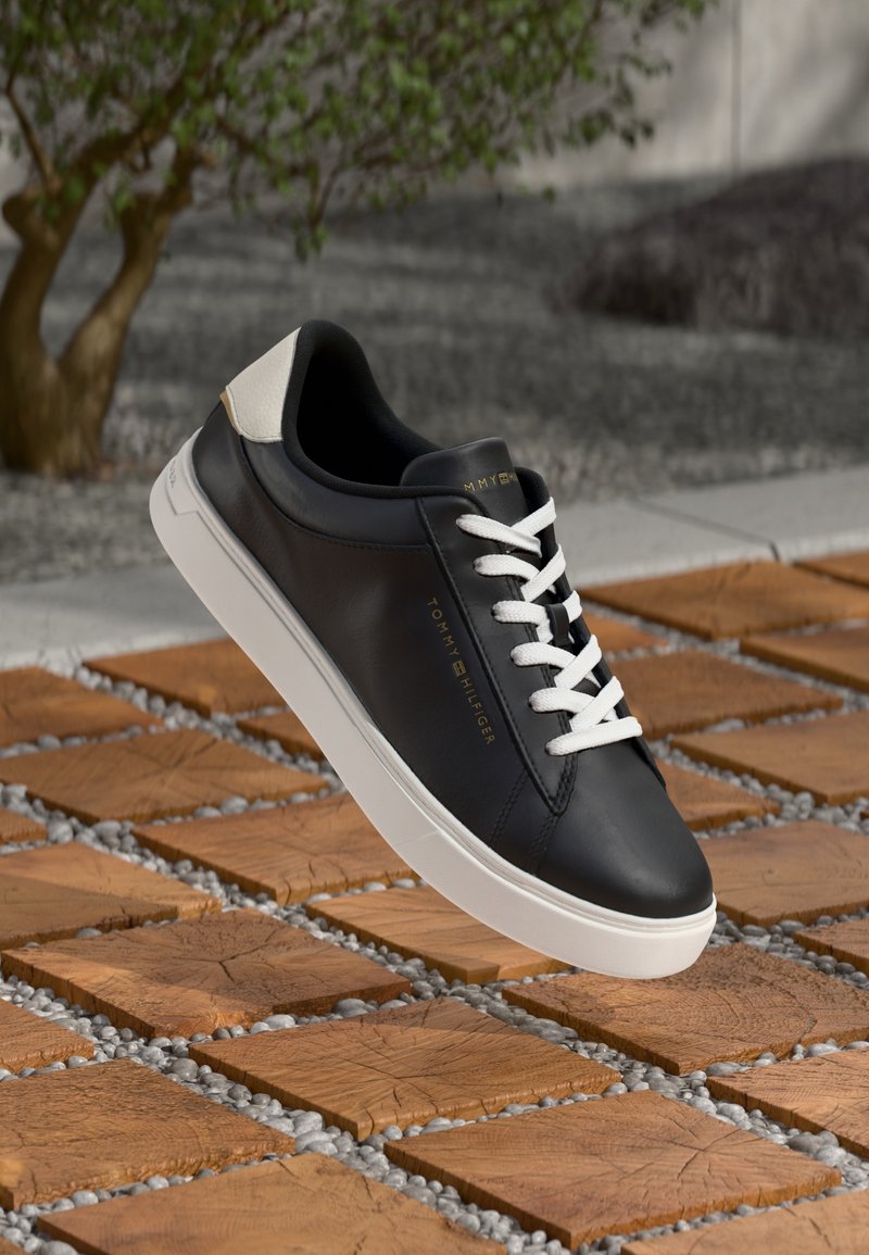 Baskets en cuir noir avec lacets et semelle blancs, marque "Tommy Hilfiger", présentées sur un sol extérieur en carreaux de bois et galets.