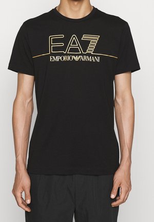 Mężczyzna w czarnej koszulce z złotym napisem i logo "EA7 Emporio Armani" na piersi, stojący na neutralnym tle.