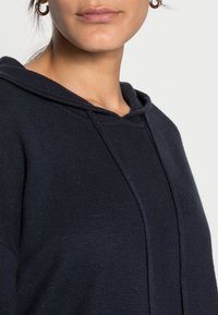 Mörkblå huvtröja med texturerat stickat material, med dragsko i huooden och minimala sömnadsdetaljer. Storlek och passform är inte specificerade.