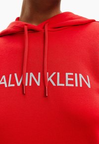 Sweat à capuche rouge avec cordons, d'une texture douce, et un logo blanc audacieux "ALVIN KLEIN" affiché en évidence sur le devant.