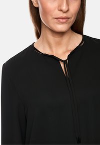 Schwarze Bluse mit V-Ausschnitt und Bindeband, gefertigt aus glattem Stoff. Das Design enthält keine sichtbaren Muster oder Beschläge.