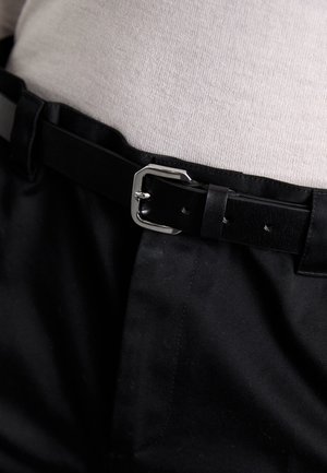 Cintura in pelle nera con fibbia rettangolare argentata, indossata sopra un tessuto grigio chiaro con trama fine, abbinata a pantaloni neri.