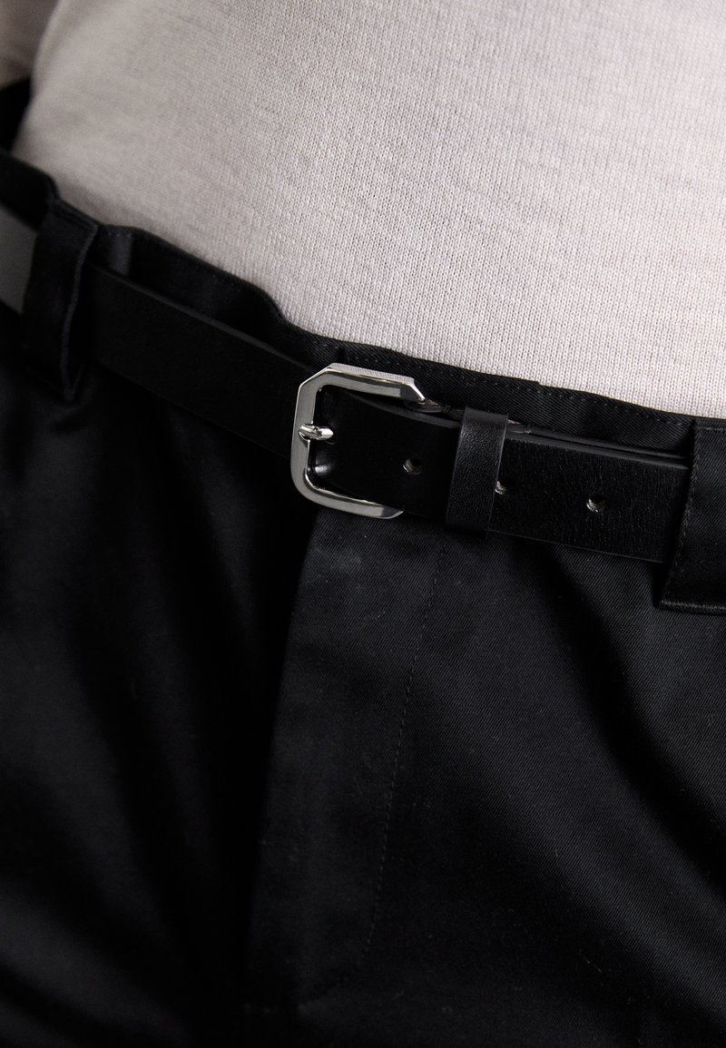 Curea din piele neagră, cu o cataramă rectangulară argintie, purtată peste un material textil gri deschis cu textură fină, asortată cu pantaloni negri.