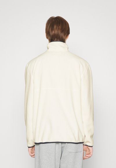 Fleecová bunda v off-white farbe s vysokým golierom, hladkou textúrou a kontrastným tmavým lemom. Disponuje uvoľneným strihom a bez viditeľných log.