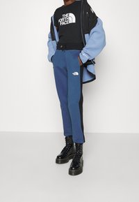 Blå fleecejacka över en svart t-shirt med logotyp, kombinerad med marinblå sweatpants med svarta sidopaneler och svarta snörboots.