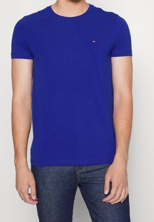 Man wearing a fitted royal blue short-sleeve t-shirt with small red, white, and navy logo on the left chest, paired with dark blue jeans.
