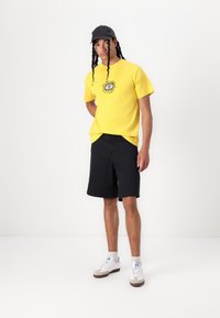 Žluté bavlněné tričko s grafickým designem, spárované s černými šortkami a bílými teniskami s šedými detaily. Černá čepice doplňuje celkový outfit.