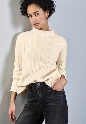 Cream ribbed sweater with high neckline and long sleeves, paired with dark gray jeans. The outfit features a relaxed fit and minimal detailing.