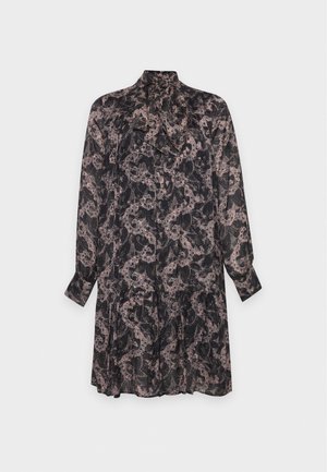 Un mannequin se tient devant un fond gris uni, vêtu d’une robe sombre à fleurs, à manches longues et avec un nœud au col, ainsi que de sandales noires.