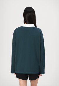 Tmavý teal dlhý rukáv košeľa s bielym golierom, s uvoľneným strihom a mäkkou tkaninou. Nosené s čiernymi šortkami.