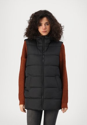 Gilet matelassé noir avec fermeture éclair, capuche et poches avant. Présente un design matelassé, associé à un pull texturé couleur rouille.