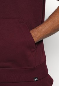 Sudadera burdeos con bolsillo canguro, puños y dobladillo acanalados. Etiqueta de marca visible en la parte inferior con acento de color contrastante.