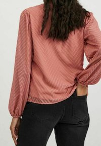 Långärmad blus i chevronmönster, i blushpink färg med puffärmar och rak nederkant, tillsammans med svarta byxor.