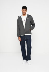 Szara, lekka kurtka z kapturem, z zamkiem błyskawicznym z przodu oraz naszywką z logo, noszona na białej koszulce i ciemnych dżinsach. Białe sneakersy.